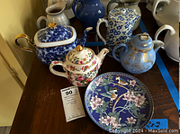 Four decorative teapots and one cloisonné plate on wooden table, showing overall lot contents including blue and white floral teapot, floral patterned teapot, blue teapot with gold dragon designs, white plain teapot, and cloisonné plate with bird and flower designs.