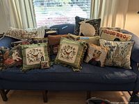 Wide view of multiple embroidered and plush throw pillows featuring animal and floral designs arranged on a blue sofa under daylight from window