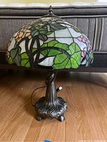 Front view of Tiffany style stained glass lamp showing colorful floral pattern and metal base on wood floor with couch in background.