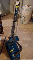 Full view of Titan upright vacuum cleaner showing blue body, hose wrapped around the handle, power cord, and base.