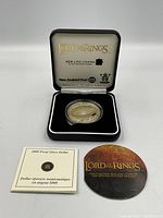 Proof silver dollar coin with 'Lord of the Rings' design in original black display box with 'Lord of the Rings' gold logo and New Zealand Post branding inside lid. Two booklets placed in front.