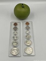 Two Canadian coin sets in plastic holders displayed with a green apple for scale, showing all coins including the large silver one-dollar coin.