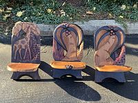 Wide shot of all 3 hand carved collapsible wooden stools with jungle animal motifs in natural wood tones and reddish accents, placed outdoors on pavement with background foliage.