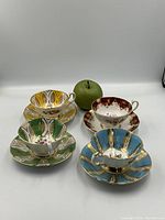 Overview of all four teacup and saucer pairs arranged with green apple for scale