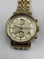Close-up view of Fossil watch face showing gold-tone stainless steel casing, champagne dial, chronograph subdials, date window, and branding.
