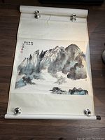 Full view of hand painted Asian scroll showing mountain landscape with clipped weights holding it flat.