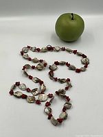 Full view of the long shell and red coral bead necklace arranged on a white surface next to a green apple for scale.