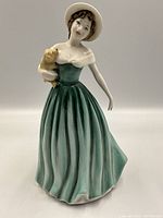 Front and side view of Royal Doulton Eleanor figurine in green dress holding small yellow dog with white hat.