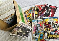 Box filled with about 100 comics with some comics laid out showing colorful front covers including Moon Knight, Deadpool, Avengers, Cable, Sabretooth, and others.