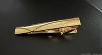 Top-down view of a gold-colored tie clip showing the textured and smooth pattern details.