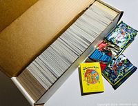 Open cardboard box showing large quantity of trading cards arranged side by side, some sealed packs visible.