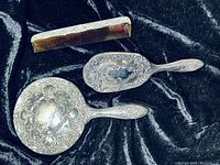 Photo showing vintage hairbrush, comb, and mirror set with ornately decorated silver-colored handles on black fabric background