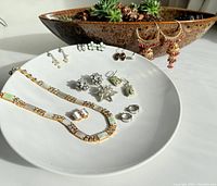 Photo displaying a jewelry set arranged on a white plate containing two gold-tone necklaces and multiple pairs of earrings with various designs including floral, rhinestone, and pearls.