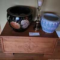 Front view of the cedar chest with black floral ceramic planter, blue and white ceramic planter, and brass lamp on top.