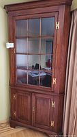 Full view of one wooden corner china cabinet showing glass-paned upper door and lower wooden double doors with brass hinges.
