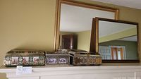Photo shows two rectangular mirrors with gold frames and three vintage German tins with embossed floral and architectural painted designs on a shelf.