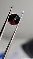 Close-up photo of a deep red Almandine garnet gemstone held by tweezers, showing the fancy diamond cut and rich color.