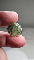 Close-up photo of round translucent mossy green prehnite cabochon with internal needle-like rutile inclusions held between thumb and index finger under natural light.