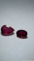 Two natural ruby gemstones displayed side by side on neutral background, showing pear and oval cuts, deep red color, some inclusions visible.