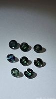 Seven round-cut blue teal green sapphires arranged in group on light background showing color variations and clarity.