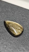 Close-up photo showing the polished pear-shaped gold sheen obsidian cabochon with strong gold flash on a textured dark surface.
