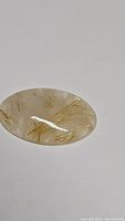 Oval cabochon shaped golden rutilated quartz gemstone lying on white surface, showing natural golden rutile needle inclusions.