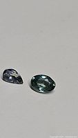 Two loose untreated Cambodian zircon gemstones, one oval teal and one pear-shaped blue-gray, on a white background.