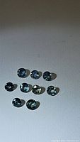 Nine natural sapphire gemstones laid on a surface showing various blended aqua blue, green, and teal tones.