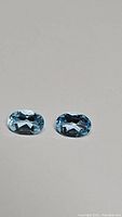 Pair of natural blue topaz oval cut gemstones shown on a neutral background. Clear oval facets and bright sky blue color visible.