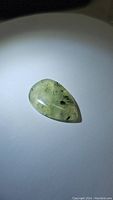 Close-up photo of a pear-shaped cabochon cut green prehnite gemstone with black rutilated needle-like inclusions, highlighting the gemstone's texture and inclusions.
