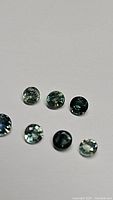 Top-down photo showing seven round cut sapphires in shades of teal green and blue on a plain background.
