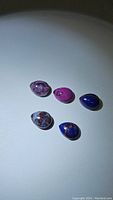 Five pear-shaped cabochon stones of varying colors and patterns including purple copper turquoise under white light on white surface.