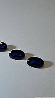 Three loose royal blue oval cut sapphires arranged in a row on a white textured surface under soft light showing deep blue color and transparency.