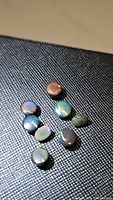 Nine small untreated Ethiopian black opal gemstones on black textured surface displaying medium flashes of red, green, and blue colors under light.