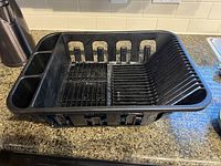 Black plastic dish rack with multiple compartments for plates, cups, and utensils on a kitchen countertop.