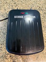 Closed view of the black George Foreman sandwich maker with brand logo and indicator lights on top.