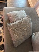 Two decorative square sofa pillows with leaf pattern placed on a gray sofa seat, showing the pattern and piping detail.