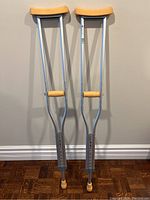 Full view of two aluminum forearm crutches with beige padded supports standing against a wall on hardwood floor.