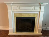 Front view of white electric fireplace with mantel and black glass doors, showing decorative molding and beige stone surround.