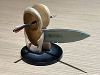 Photo showing front and side angles of painted resin sculpture of ears pierced by arrow and holding blade on black base.