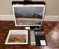 Black portfolio with large photo print of a mountainous landscape with a rainbow, framed autumn cityscape photo, and Canson Infinity printer paper box on hardwood floor