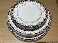 Stack of white Wedgwood Medici lunch plates with detailed brown and dark blue shell-pattern border, viewed from above.