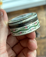 Close-up angled photo showing the round pill box with guilloche enamel floral decoration and pink and green trim held in hand, highlighting detailed painted roses.