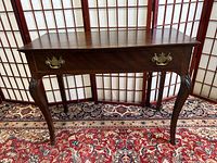 Front view of antique wooden side table showing one dovetail drawer with brass handles and bow legs on a patterned rug against a folding screen.