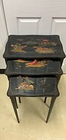 Stacked view of three nested black lacquered Asian nesting tables with hand-painted detailed designs including pagodas, birds, and foliage on top surfaces.