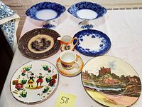 Full view of the ceramic dishware lot including cake plates, transferware plates, colorful folk art plate, and small pitcher and cup.