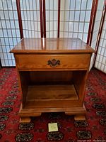 Front view of the Ethan Allen wooden side table showing one drawer with brass handle and open shelf below. Table surface and drawer have random scratches. Furnished with carved feet and medium brown finish.