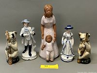 Five porcelain and ceramic figurines arranged in a row on a white surface: two koalas holding logs on each end, a man playing a stringed instrument, a woman dressed in a white dress with blue trim and hat, and a taller woman with a smaller child figurine in front.