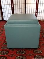 Front view of the green vinyl padded storage ottoman showing its square shape and reversible top.
