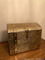Front view of the brass tone metal treasure chest with domed lid, faux clasp, and studded metal straps.